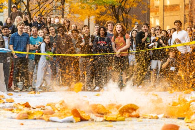 Photo: An action shot of a pumpkin smashing into the ground. Pieces of pumpkin fly from the impact as people standing a distance away react with joy at the smashing.