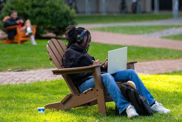 Photo: A young Black woman with long braids tied up in a half up hairstyle and wearing large black Beats headphones, a black fluffy top, and jeans, sits in a large brown lawn chair as she studies on an open laptop, phone, and tablet. She sits in a large lawn on a sunny day.