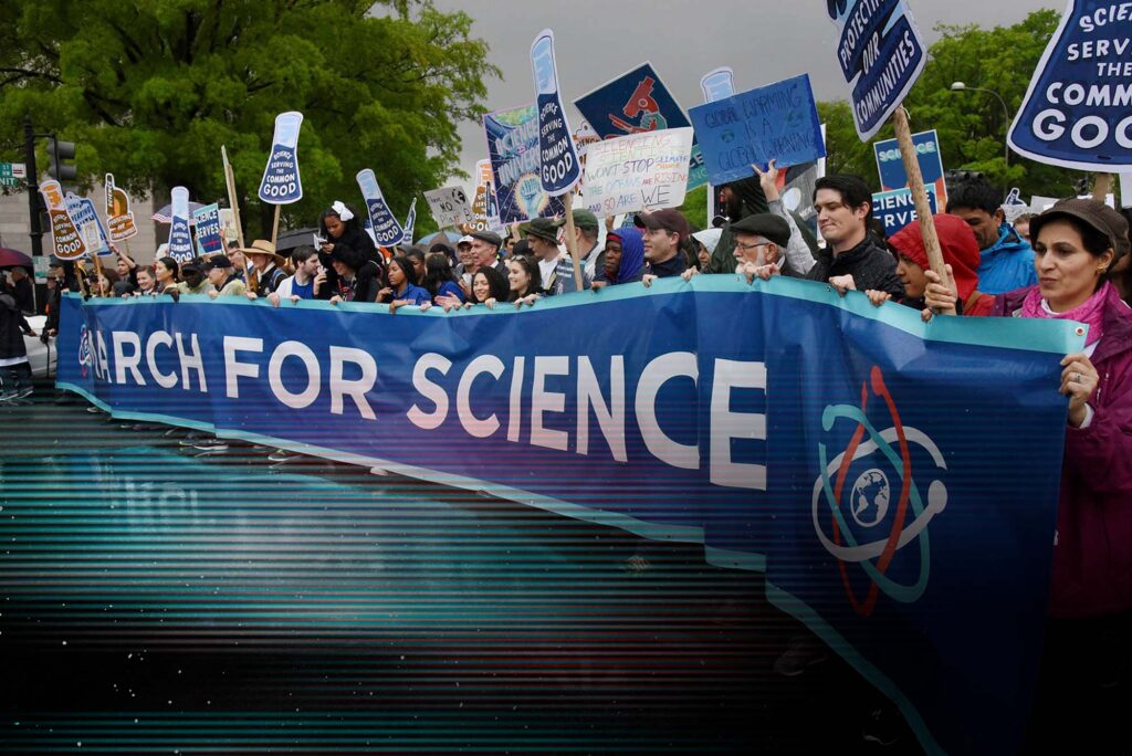 Photo: Thousands of scientists and their supporters join the March for Science in Washington, D.C. A crowd of people hold signs and chant on a rainy, gloomy day. A row of people int he front hold a large blue banner across them that reads "March for Science".