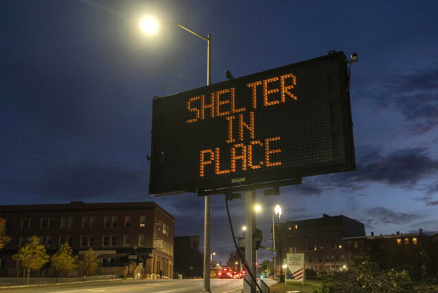 Photo: A large black road sign reads "Shelter in Place" in LED letters on a dark evening in Maine. Background sign shows an empty street.