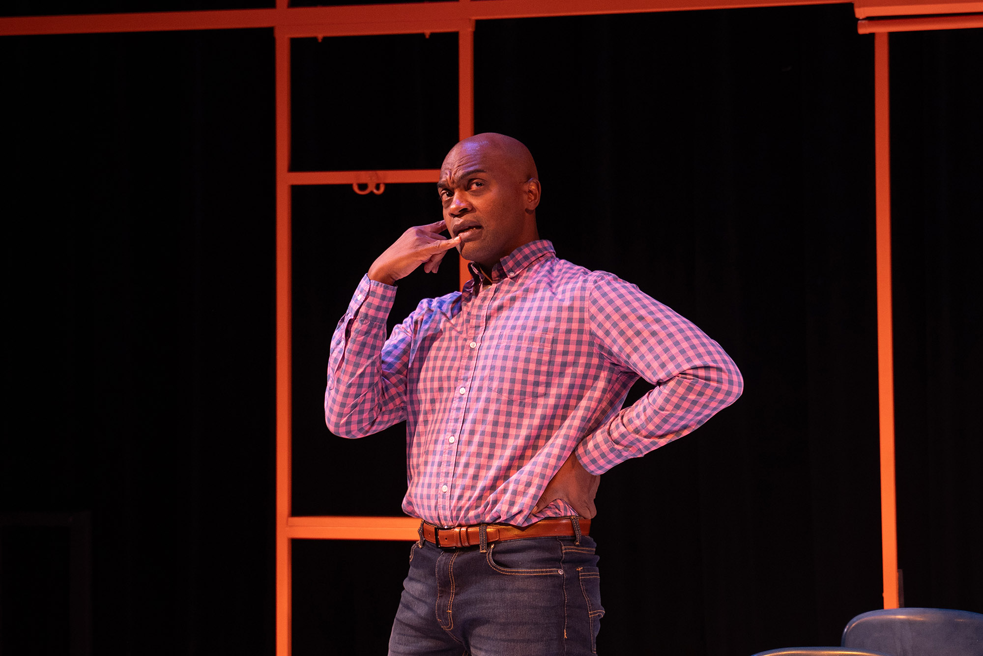 Photo: Maurice Parent, a tall, bald Black man wearing a long-sleeved burgundy, striped collared shirt and jeans, holds one hand to his hip. His other hand is formed into the "phone" motion as he mimes talking into a phone on a stage.