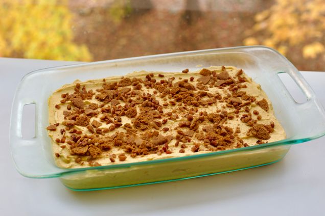 Photo: A large glass dish filled with a light orange pumpkin mousse. The top of the dessert is sprinkled with small, brown gingersnap cookie chunks. Dessert is displayed on a white table in front of window. Scene outside is a fall scene of leaves changing color.