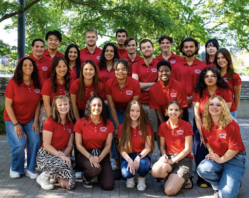 (Photo Credits): This is a photo of Boston University Student Employment Office student workers. They are posed in four different rows. In the back three rows, the students are standing and smiling. In the front row, the students are kneeling down. Each student is wearing a red polo that has the Boston University logo in the right top corner. In the back of the photo, there is a green tree and they are standing outside on stone. The students are wearing various different bottoms. Most are wearing jeans but one student is wearing brown shorts in the front, the female to the left of her is wearing brown pants, and to the left of her, the individual is wearing a brown zebra print maxi skirt.