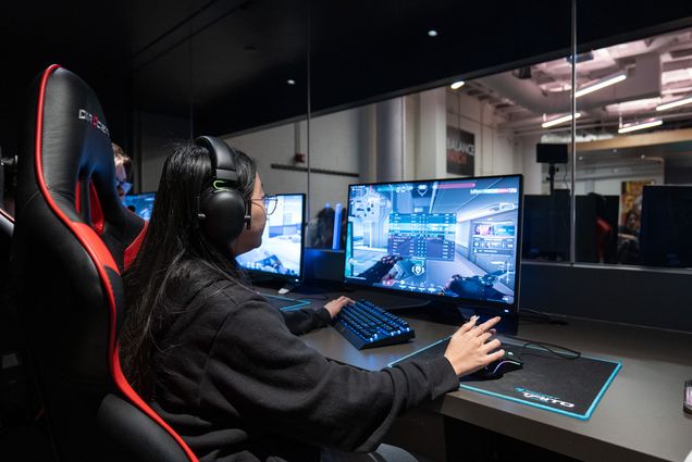 Photo: A young BU student plays an online game at Balance Patch, the on-campus video game lounge. She sits in a gaming chair, looking at a computer screen and plays with a keyboard and mouse.