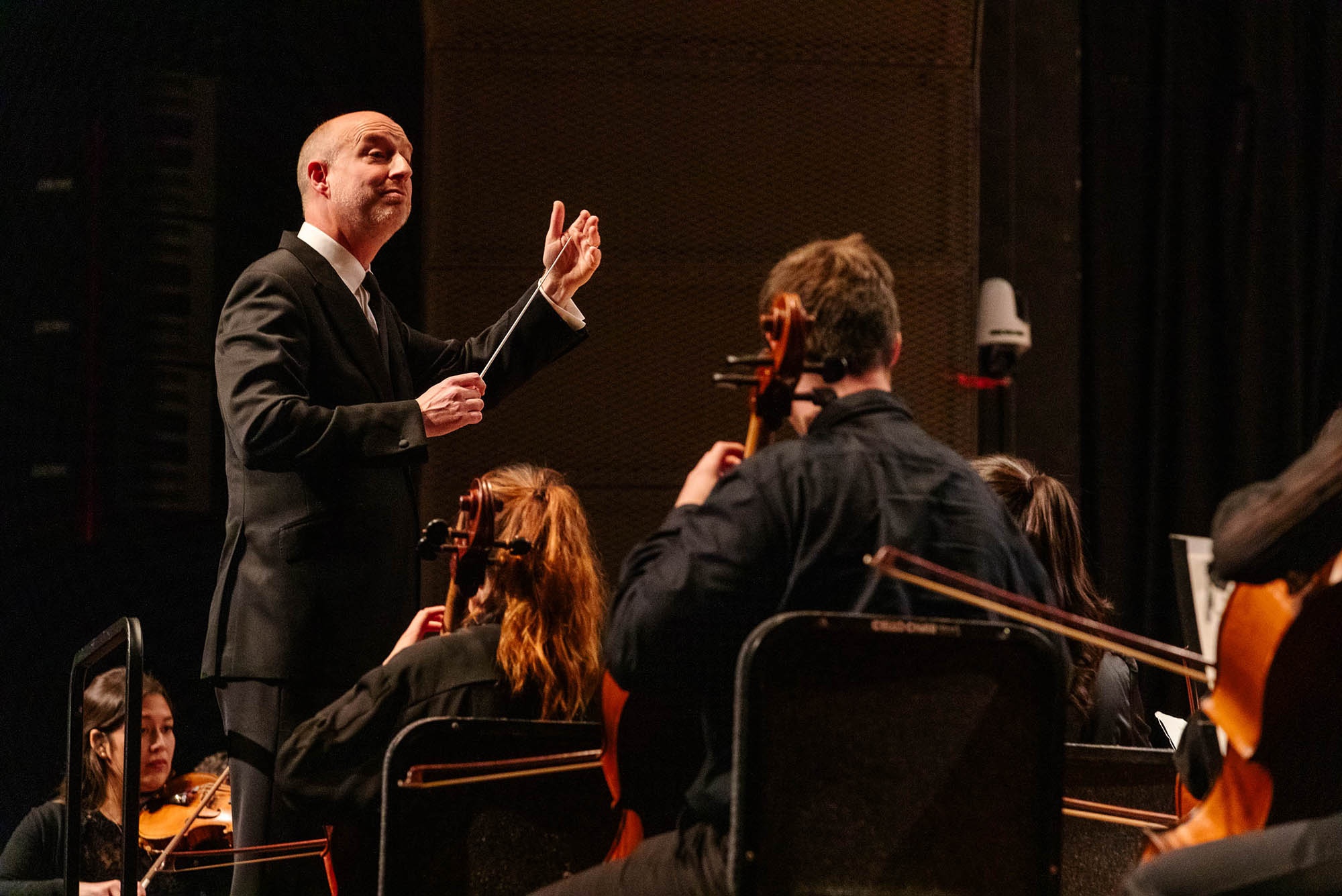 Photo: James Burton, a bald white man wearing a black suit ensemble and holding a conducting wand, as he directs student musicians wearing all black and seated playing various instruments in front of him.