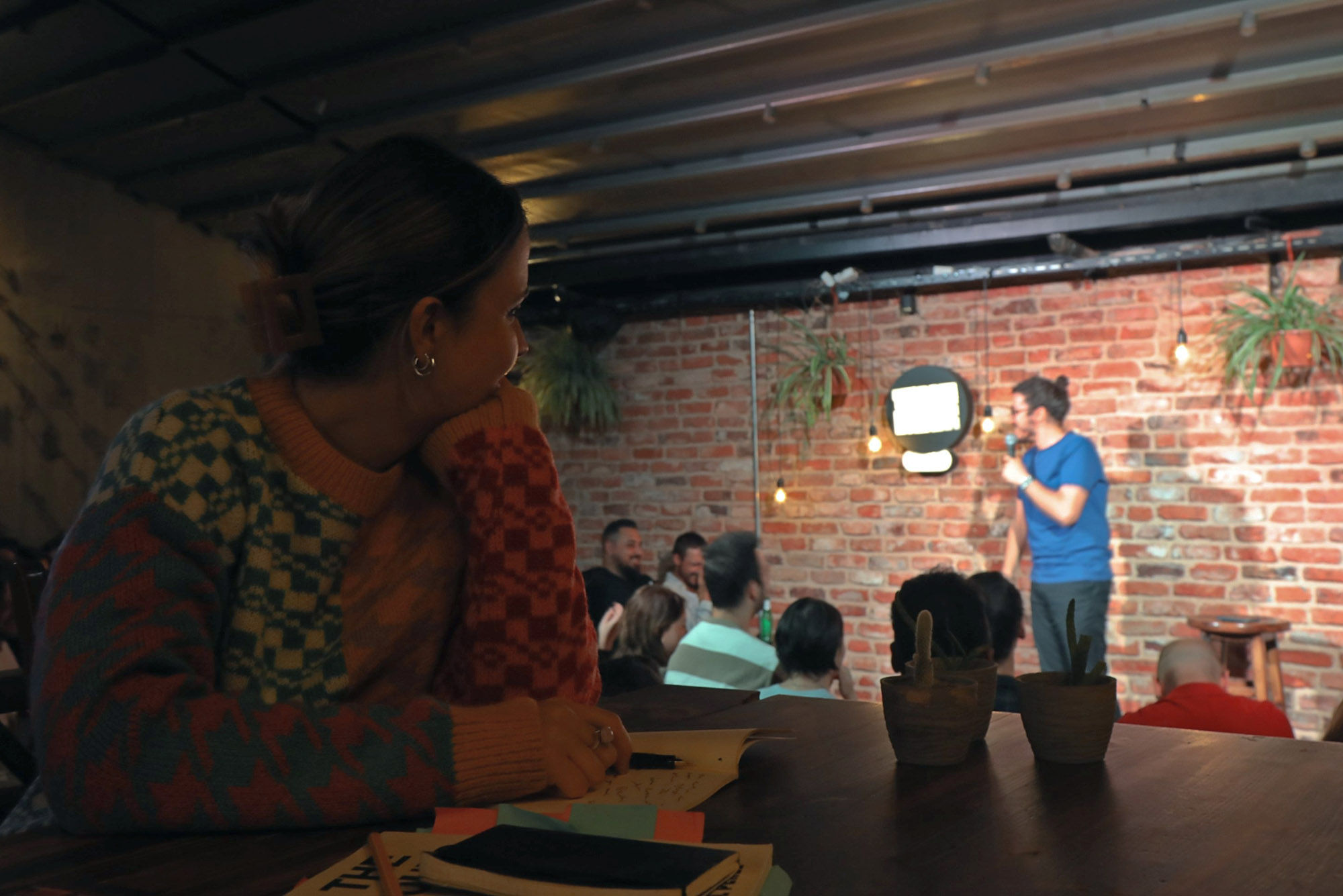 Photo: In the foreground, a young woman wearing a colorful sweater and taking notes in a notebook looks to her left towards a comedian on stage, telling jokes under a spotlight to a small audience in front of them.