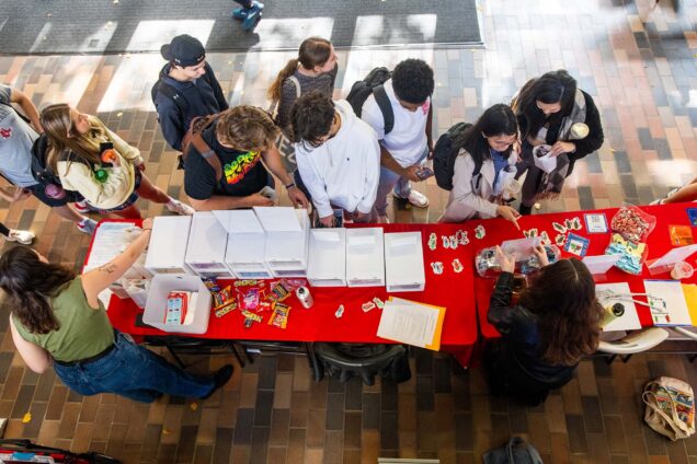 Photo: Overhead shot of students gathering around the Condom Fairy Pop-up! event in the BU GSU. A group of young adults look and interact with a red table full of sexual safety items and educational material.