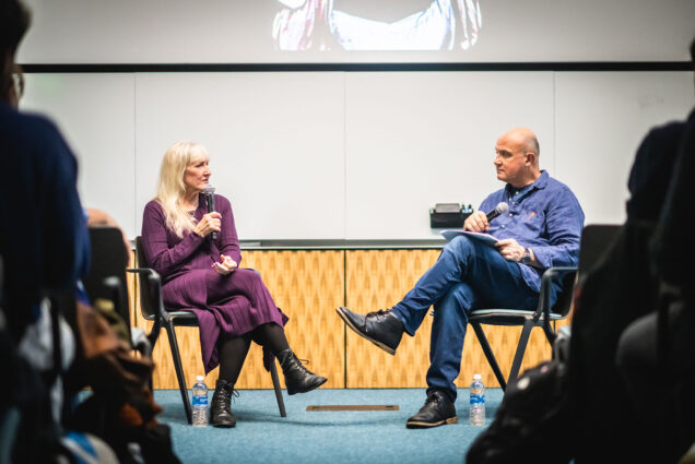 Photo: Carol Guzy, a white woman with long blonde hair and wearing a purple dress and black boots, talks into a microphone as she sits in black chair in front of a seated audience. She speaks to a moderator to her right. Moderator is Greg Marinovich, a white bald man wearing a blue shirt and jeans. They sit in front of a projector displaying some of Guzy's photos. Scene is shown down the aisle between the seated audience.