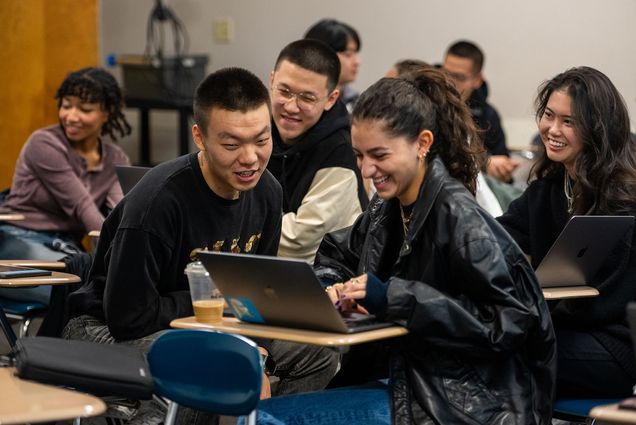 Photo: a classroom of students seated at desks smile as they work on partner assignments together. They share laptops as they work closely.