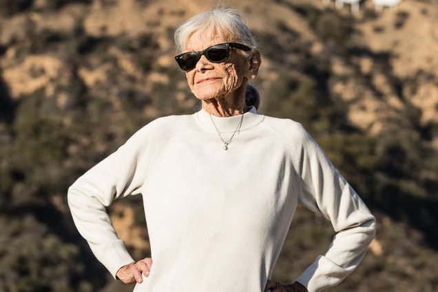 Photo: Jo Farkas, an elderly white woman wearing sunglasses, a white long-sleeved sweater, and jeans, smiles and poses with hands on hips on a sunny day. Behind her, desert hills and the white Hollywood sign can be seen in the distance.