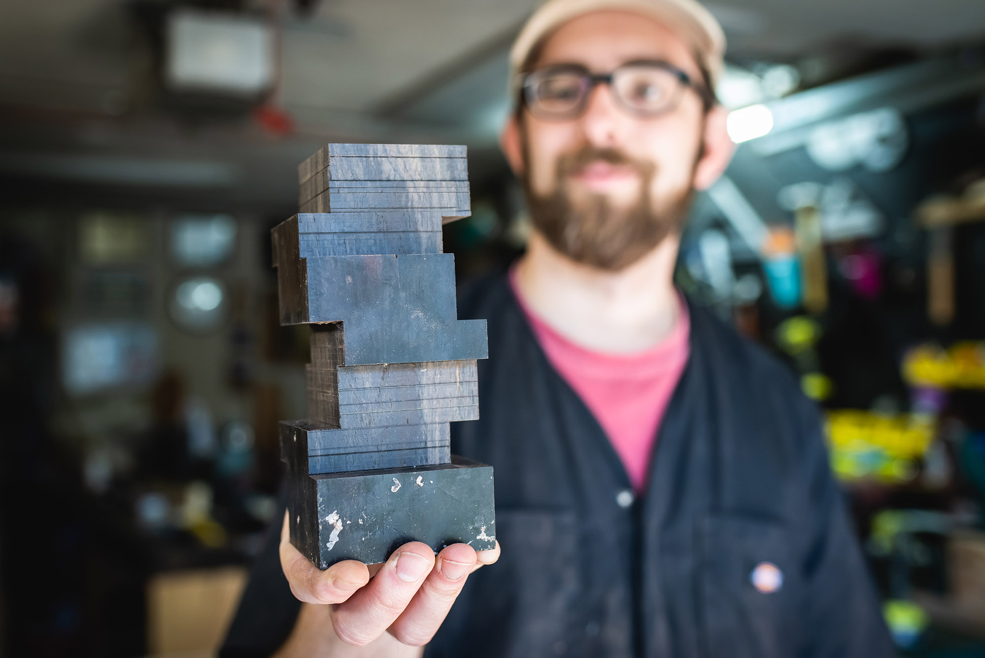 Photo: Micah Sieber, a white man with brown beard and mustache and wearing a tan cap, red-pink shirt, black collared shirt, and black pants, smiles and holds up a small black, model building. The building is in the shape of the BU CDS building, a Jenga-like building with smooth sides.