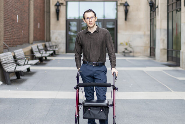 Photo: A young white man wearing glasses, a brown, long sleeved collared shirt and navy blue slacks, stands and poses with the aid of a mobility device. He poses in front of BU's College of Fine Arts.