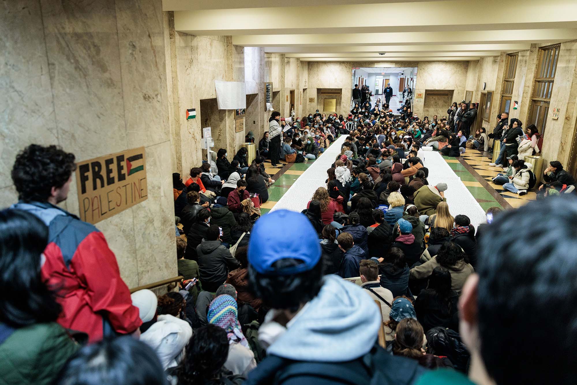 Photo: A large crowd of students sit in a large Performance Center entrance hall in silence. some are masked and some now. In the foreground to the left, a sign posted to a wall reads "Free Palestine". Two large rows of scrolls can be seen in the center of the crowd.
