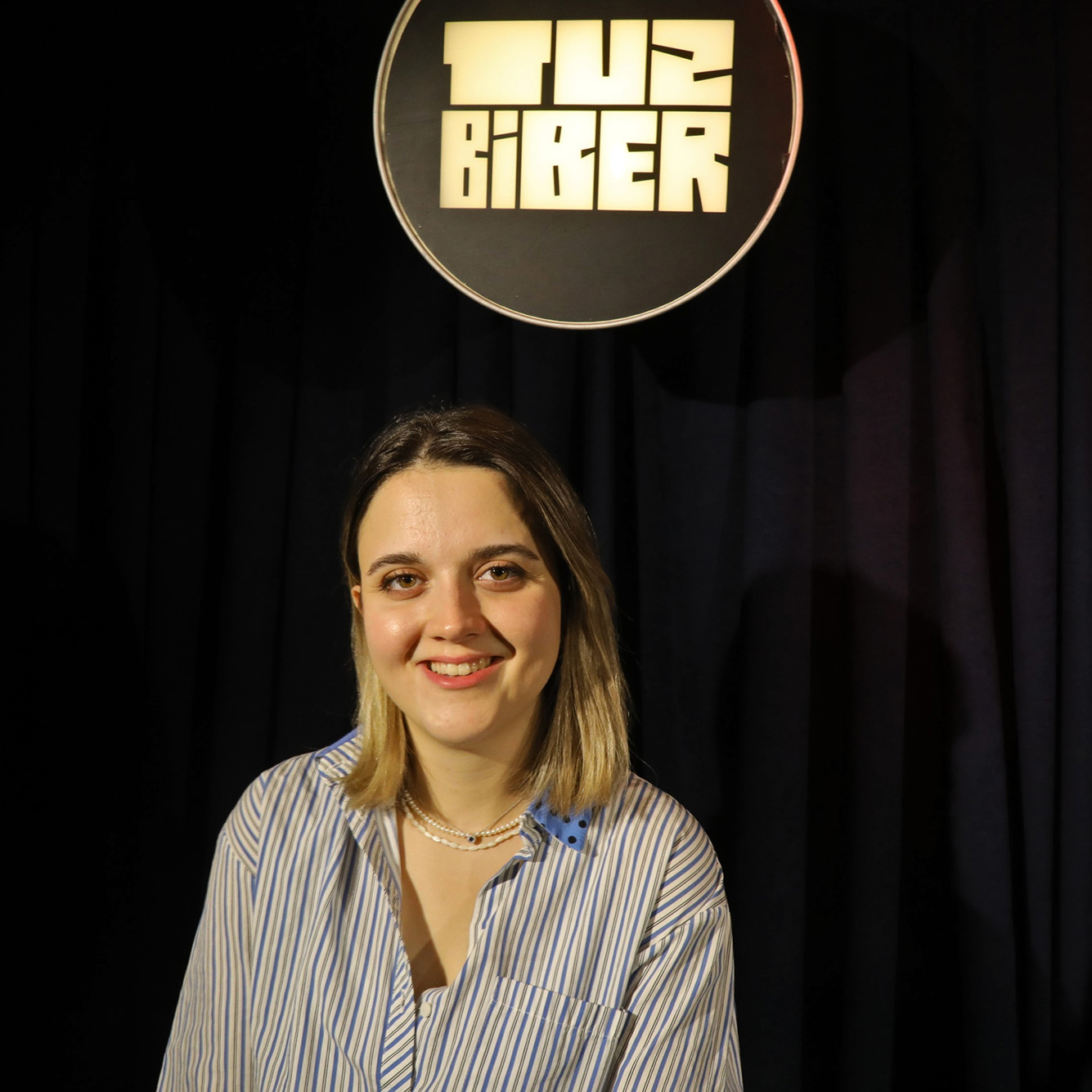 Photo: Sümeyra Güneş, a white woman with short brown and blonde hair and wearing a blue and white striped collared shirt, smiles and poses in front of a black curtain with a sign that reads the comedy club's name in Turkish.