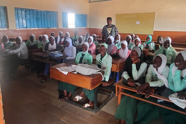 Photo: A classroom full of individuals smiling at the camera. The teacher stands in the back. smiling as well.