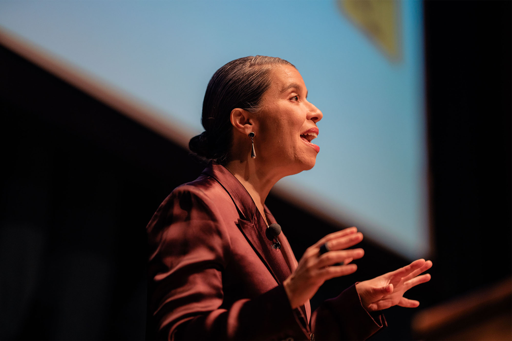 Photo: closeup of a woman in a suit lecturing in front of a dropdown screen 