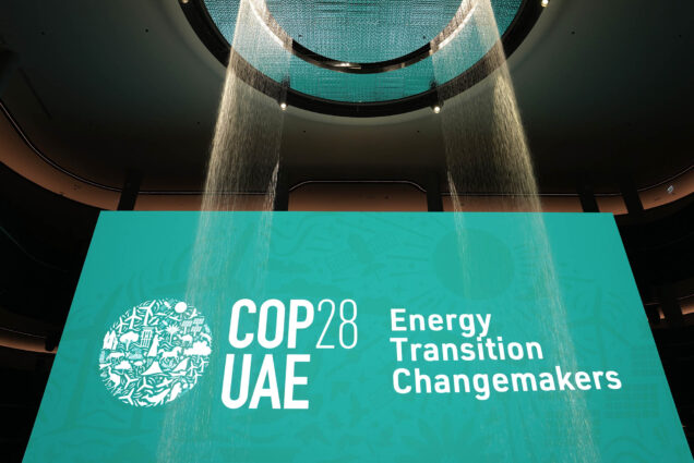 Photo: A large teal sign reads "COP28 UAE: Energy Transition Changemakers". Streams of lit up water streams are shown in front of the bright sign.