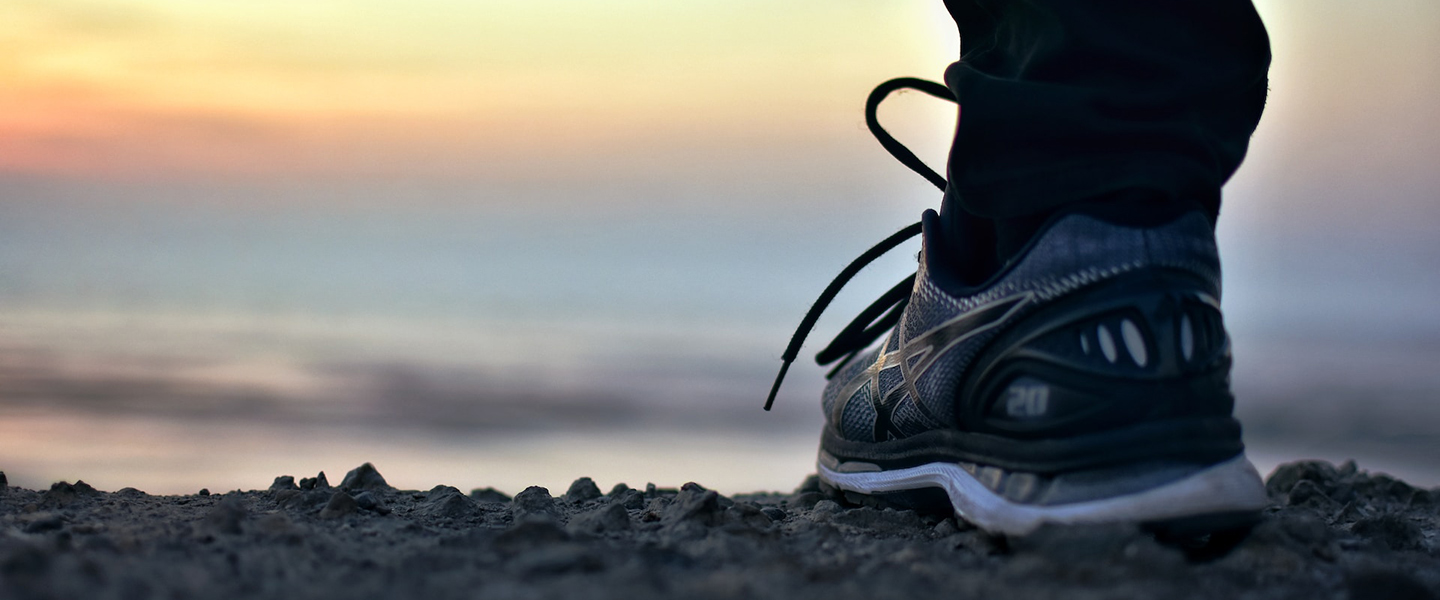 Photo: Closeup photo shows a person's foot wearing a sneaker. They stand on a dirt path and look towards a sunrise.