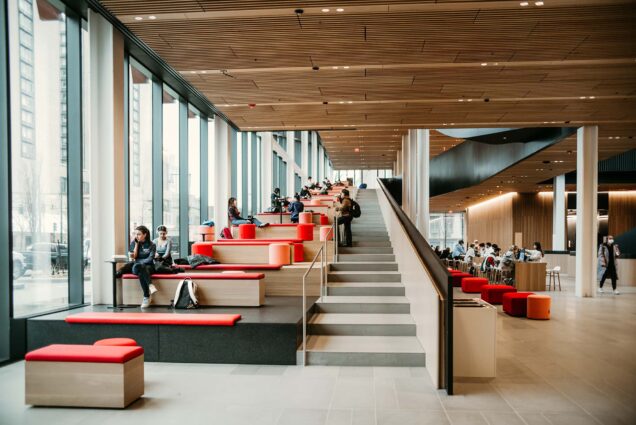 Photo: Interior of Boston University’s Center for Computing & Data Sciences. Large walkway staircase i shown. Next to the staircase, a multifunctional space for studying, hanging out, or hosting events is shown with modular black, wood, and red seating areas. Two the far left are large windows letting in lots of natural light. A vast wooden ceiling tops off the area.