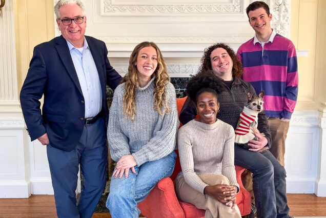 Photo: A diverse group of people pose in front of an unlit fireplace in a brightly sunlit room. In the center sits a young Black woman wearing an oatmeal-colored sweater and khaki pants. Sitting on the armchair to her right is a white man with curly hair. A small dog sits in his lap. To her left, sitting on the other armrest is a young white woman wearing a grey sweater and jeans. Behind her stands an older white man wearing glasses, a light blue collared shirt, navy blue blazer, and jeans. To the far right stands a young white man wearing a pink and blue striped collared shirt. They all smile and pose together.