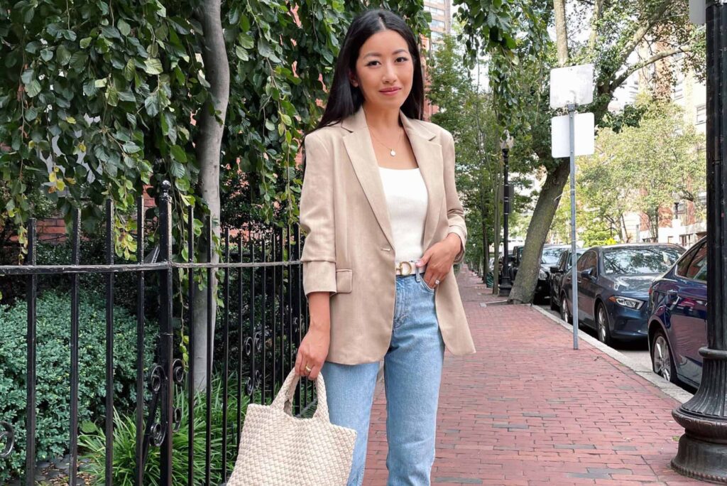 Photo: A young Asian woman with long, straight black hair smiles and poses on a quiet, red brick sidewalk. She wears a whit blouse, beige blazer, and jeans and holds a tan tote bag in her hand.