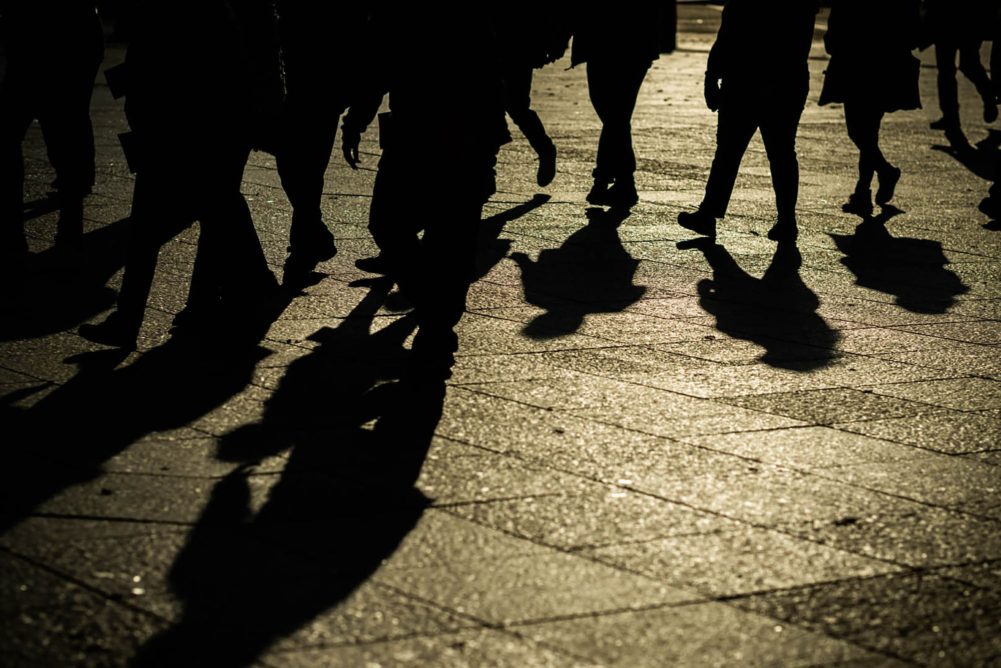 Photo: Shadowy photo shows students walking on the sidewalk of BU's campus. Photo focuses in on their legs and feet and displays everything in shadowy context.