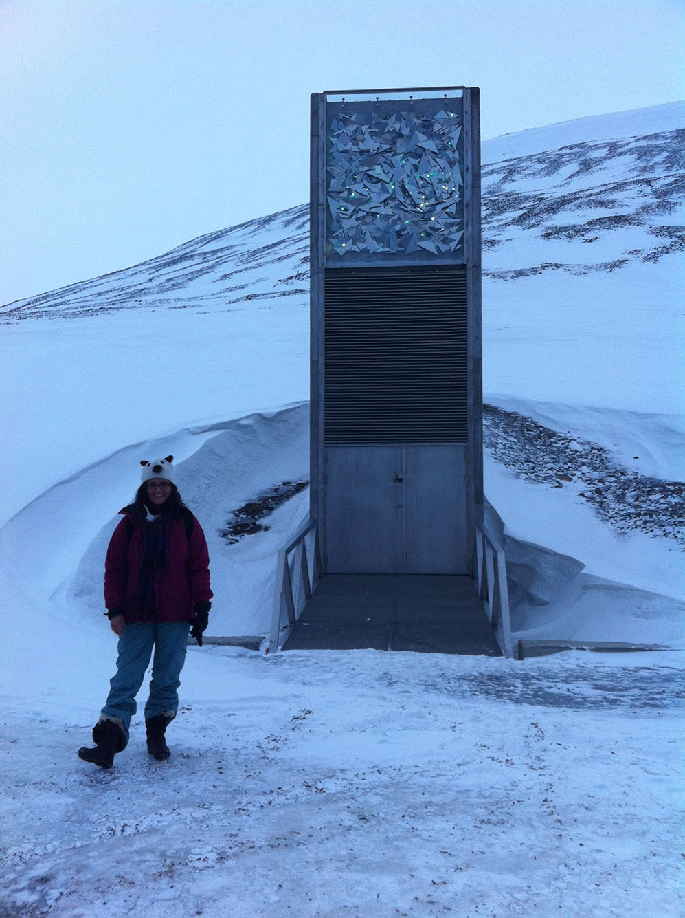 Photo: A white woman bundled up in Black winter coat, pants, hat, and boots stands in front of a large metal entrance in the middle of a snowy mountain.