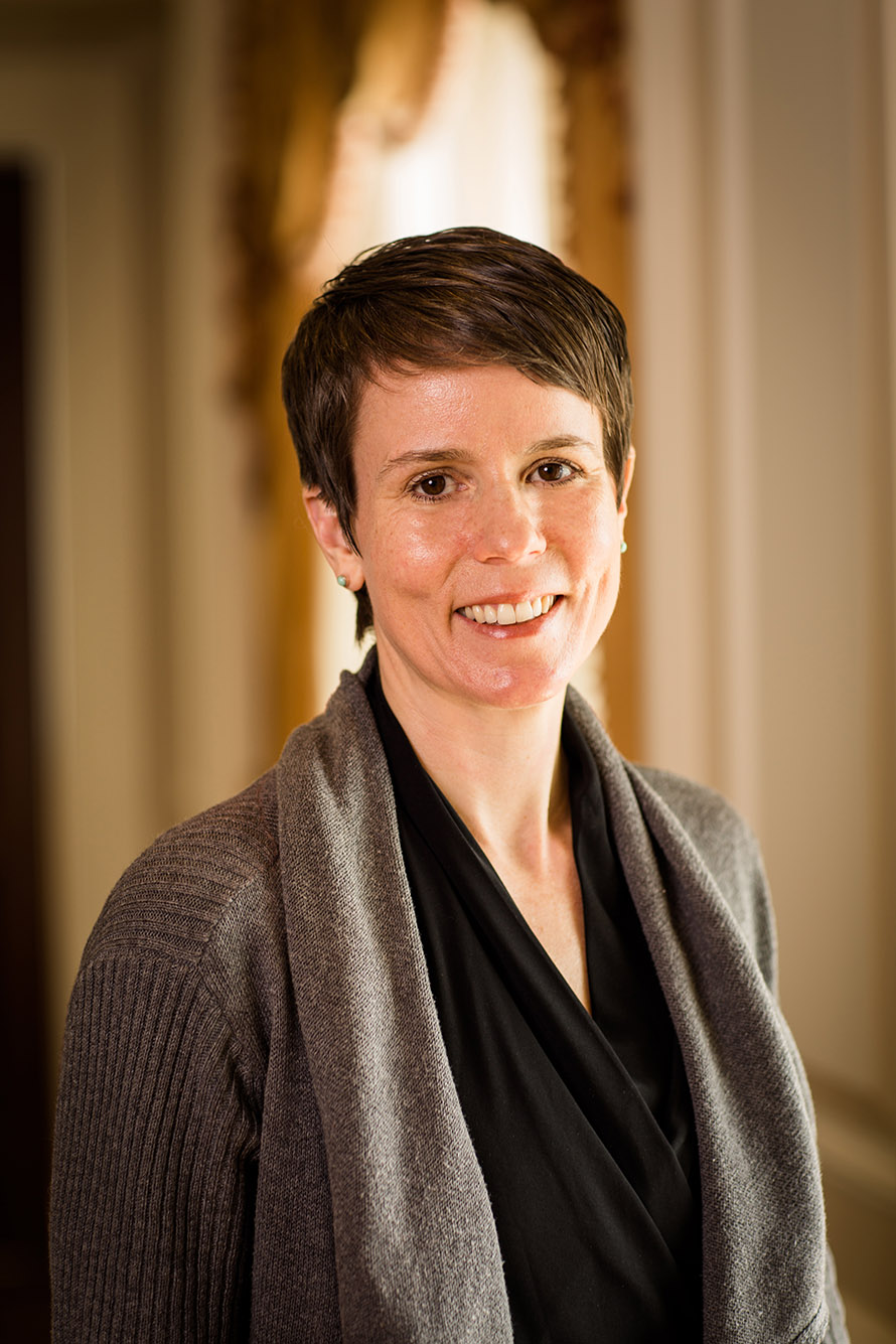 Photo: Headshot of Tracy Schroeder, a white woman with short brown hair and wearing a black blouse and brown cardigan sweater. She smiles in front of blurred office background.