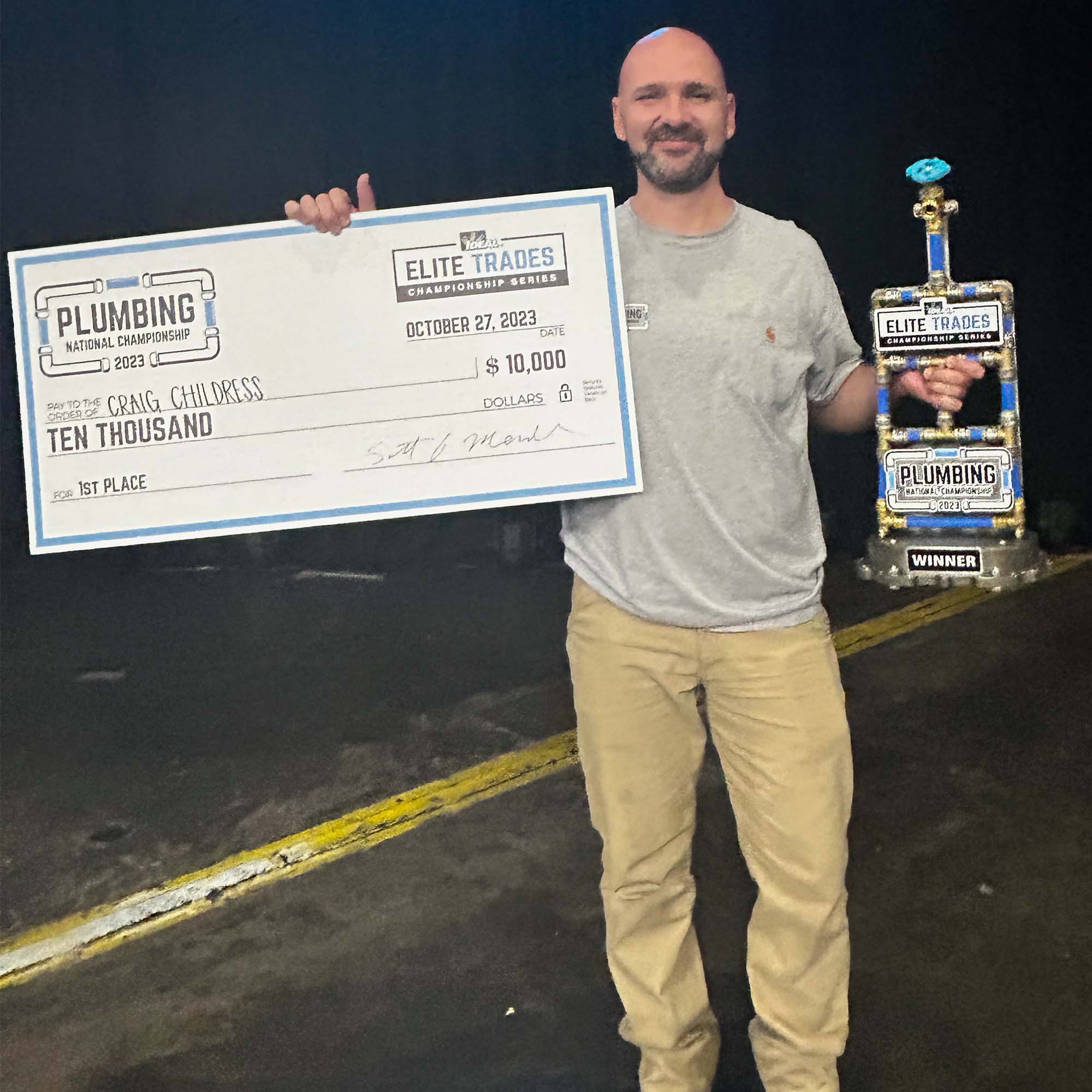Photo: Craig Childress, a white bald man wearing a grey shirt and khaki pants holds a silver trophy in his right hand and a large check for $10,000 in his left. He smiles and poses for the photo.