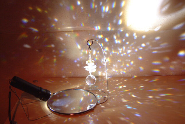 Photo: A mirror with a flash light refracting the light into scattered rainbows across the plain background. Hanging above the mirror is a dangling sun catcher, the cause of the disperse of light.