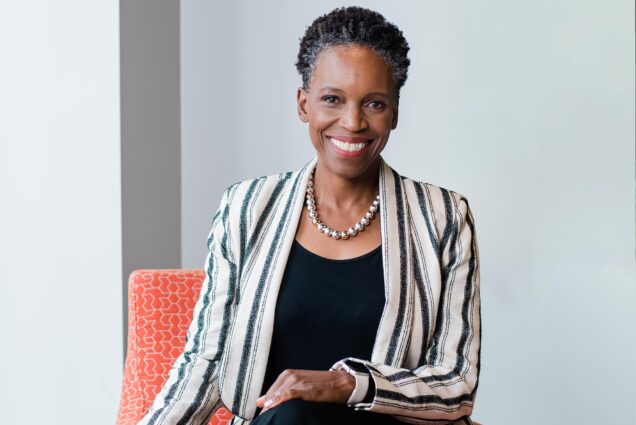 Photo: A black woman with short hair wearing a colorful striped blazer over a black shirt with a necklace, smiling for a portait in front of a gray background in a brown leather armchair