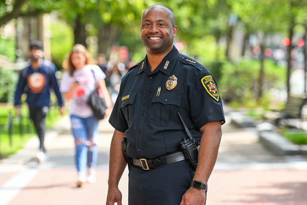 Photo: Robert Lowe, a bald black man wearing a Boston University Police Department uniform, smiles and poses on a busy sidewalk lined with greenery. Blurred pedestrians walk behind him.