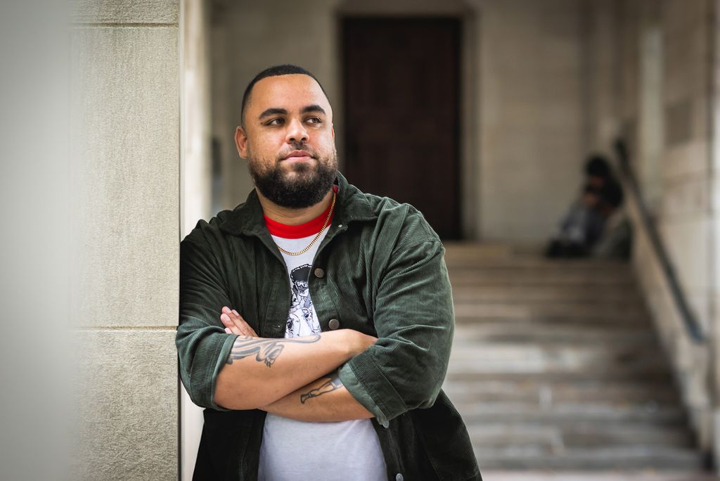 Photo: Darien Alexander Williams, a man with short, dark hair and a beard poses with his arms crossed as he leans against the wall. As he looks to the right, he wears a green button up and a white shirt with a red collar.