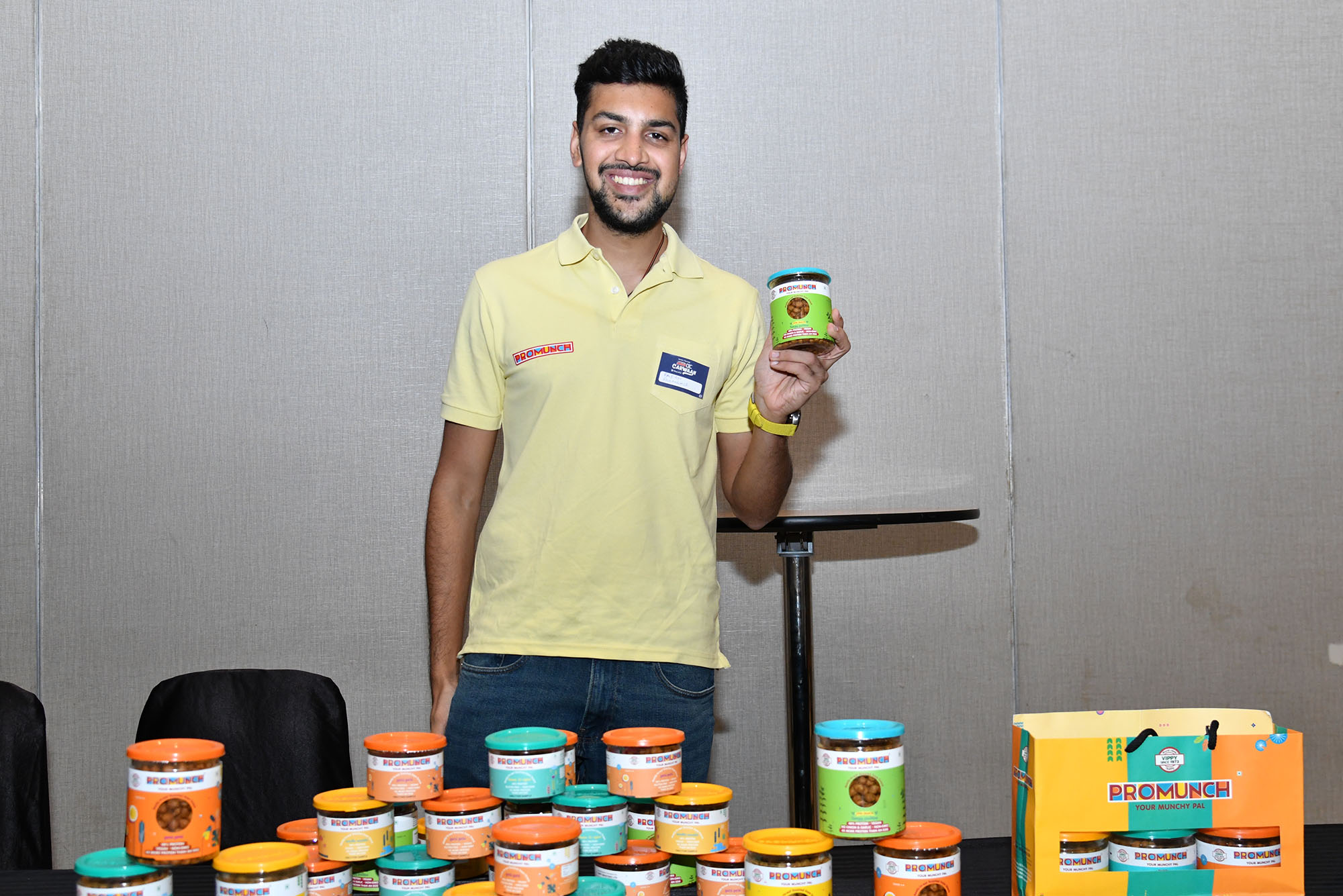 Photo: A young man poses with his product, colorful tins of vegan-friendly food, as he wears a yellow shirt and smiles for the camera.