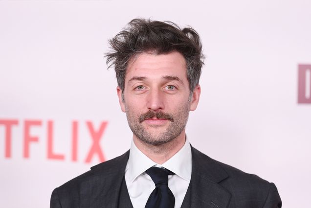 Photo: Sam Lisenco, a white man with stubble and a prominent mustache, poses for a photo on a red carpet. The picture is from the shoulders up, showing him in his black suit and black tie.