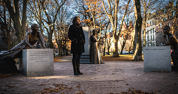 Photo: A women stands outside in the cold with three statues. There are dead trees behind her, indicating it is winter.