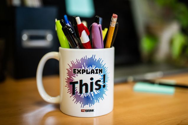 Photo: A white mug with a logo overlay reading EXPLAIN THIS! The text is featured over a burst design of purple and blue. Various pens and pencils are in the mug. The blurred background depicts office-type material.