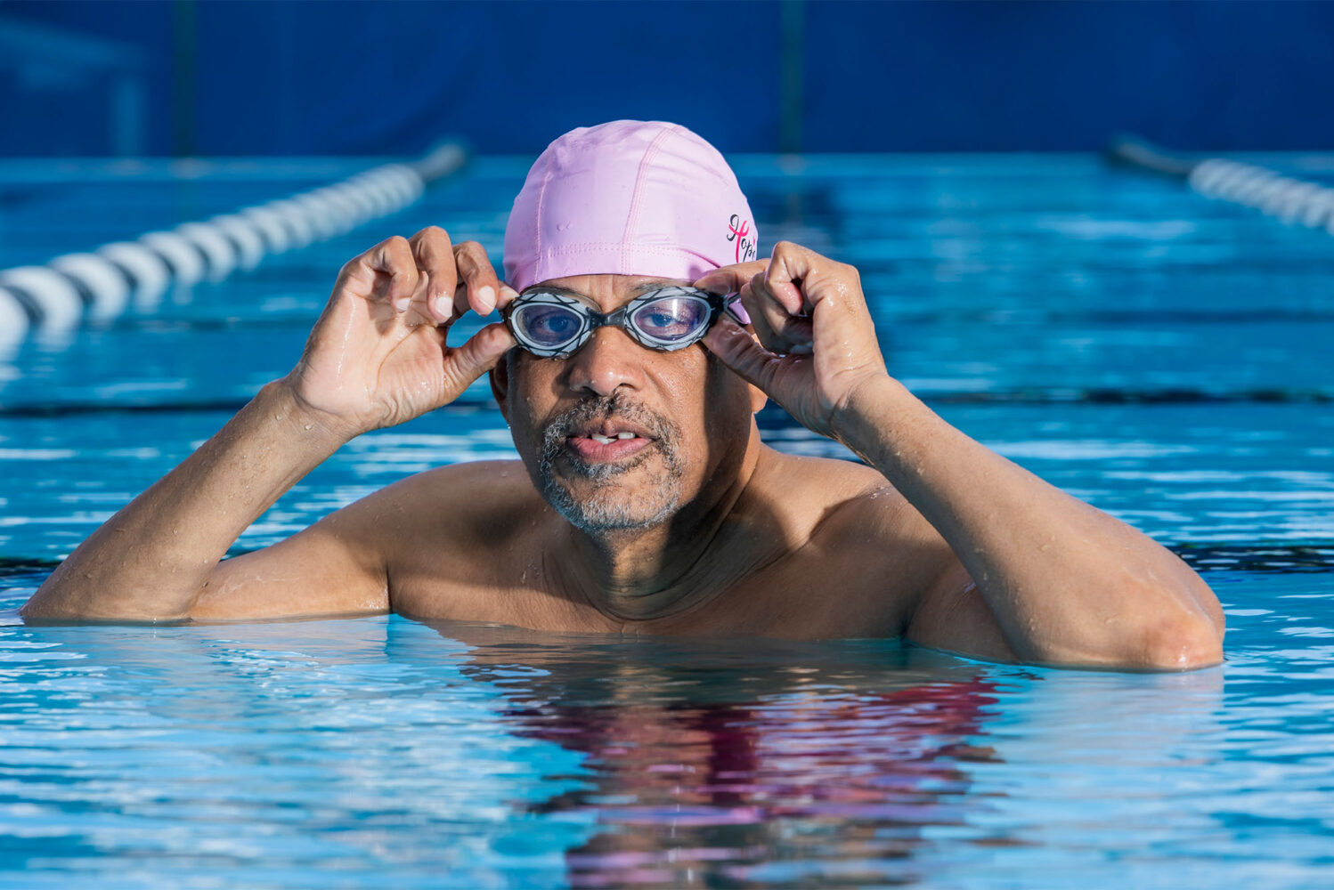 Photo: An older black man wearing goggles and a swim cap poses for a portrait in a pool