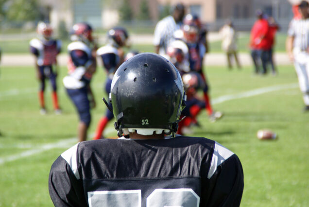 Photo: A stock photo of a kid playing American football. He is wearing a black helmet and jersey as he looks onto the field.
