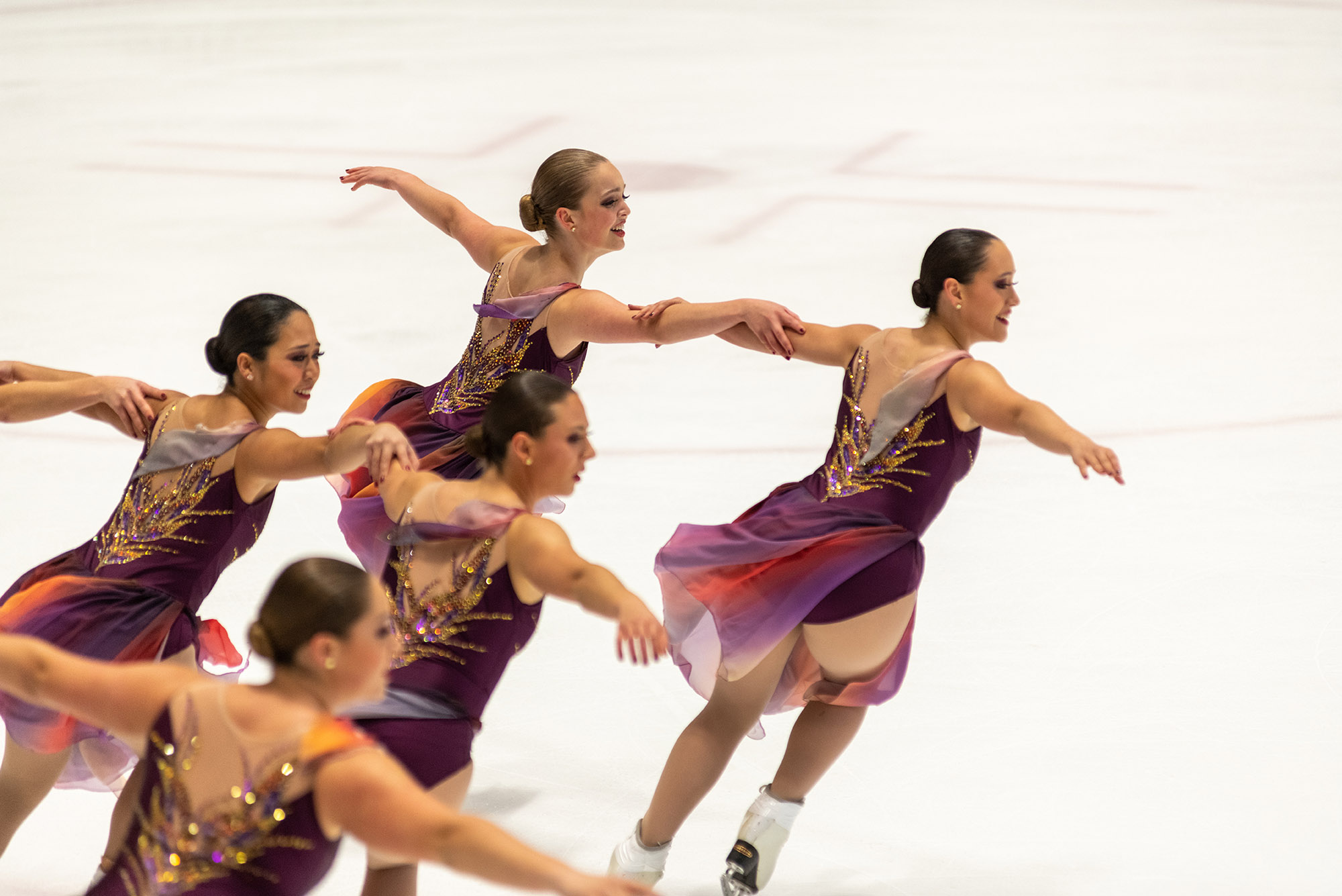 Photo: A group of figure skaters, arm in arm, skate and glide across the ice as they perform in their purple uniforms.