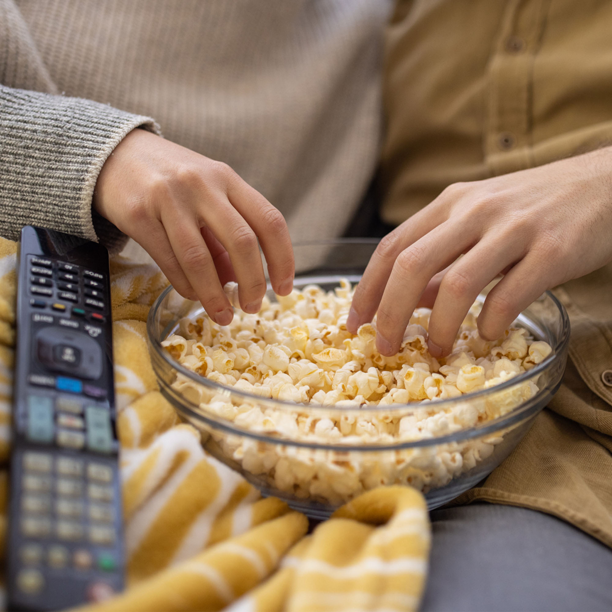 Photo: two hands reaching into a full bowl of popcorn. There is a TV remote laying next to the bowl.