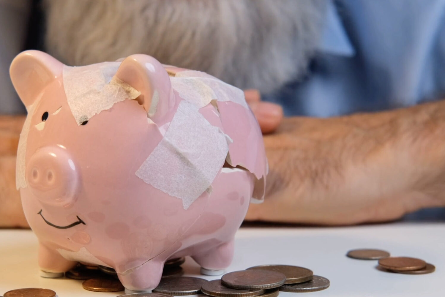 Photo: An older man with a beard sits at a table in front of a broken piggy bank. There are coins all over the table.