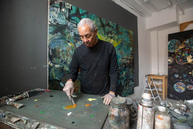 Photo: Photo: Artist Bob Freeman, a Black man with graying hair and a gray mustache, (CFA’71,’83) in his Waltham studio. The walls are used to hang his pieces, which are behind him. He is mixing paint on a slab while wearing a black turtleneck.