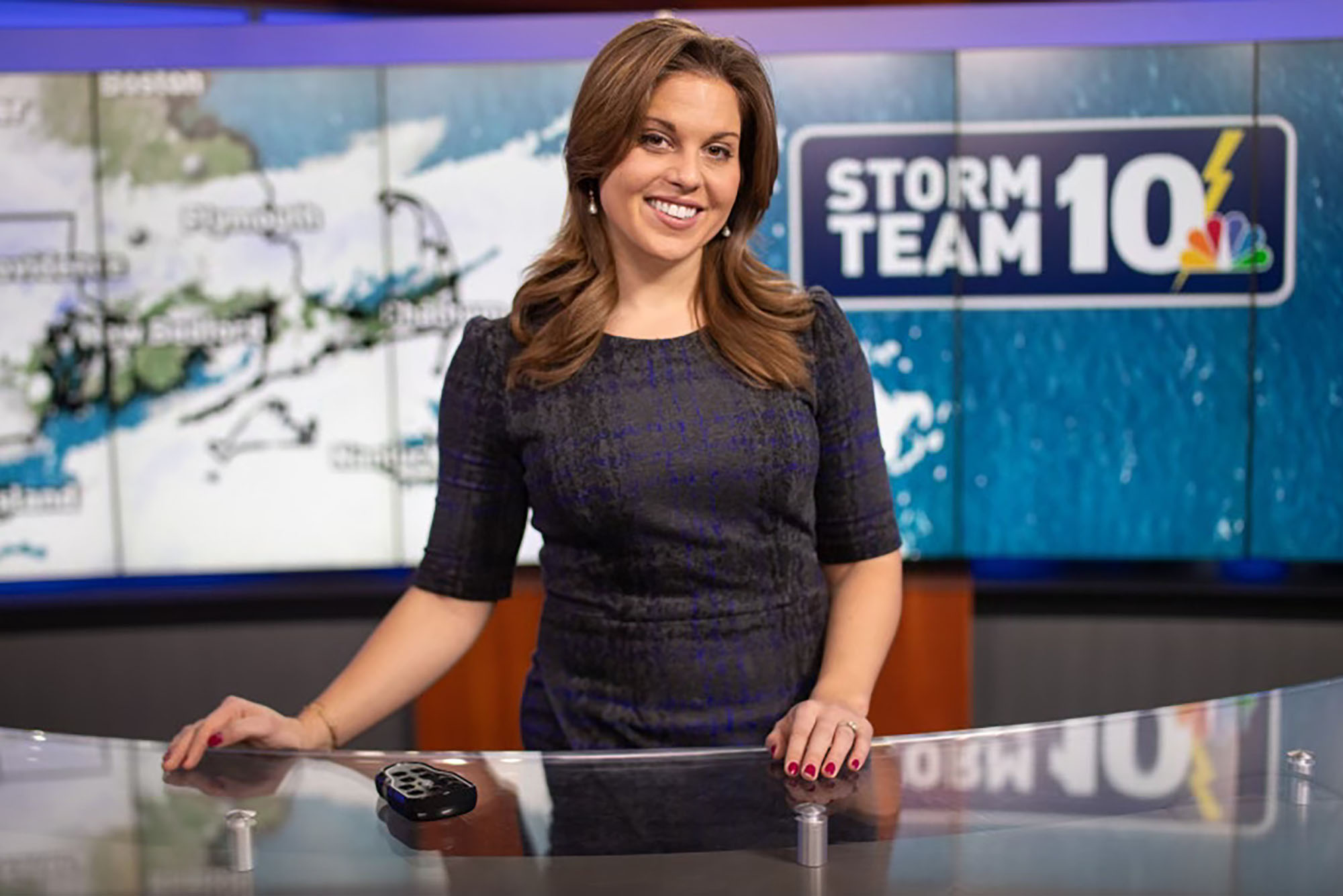 Photo: Christina Erne, a young woman with medium length brown hair and a dark dress sits at a news desk with the logo of the news station behind her left shoulder.