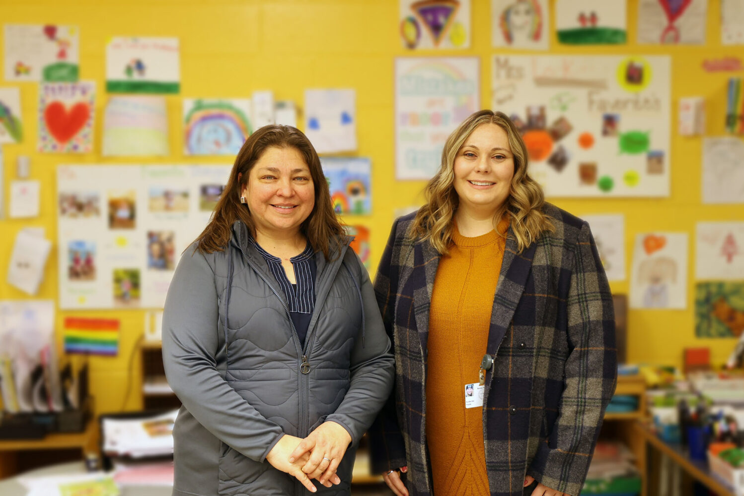 Photo: Two woman stand side by side and pose for a picture. On the left, a shorter woman with a gray cardigan and dark hair. On the right, a slightly taller woman with light hair and a plaid cardigan. They pose in a classroom setting, a yellow wall decorated with student work as the background.