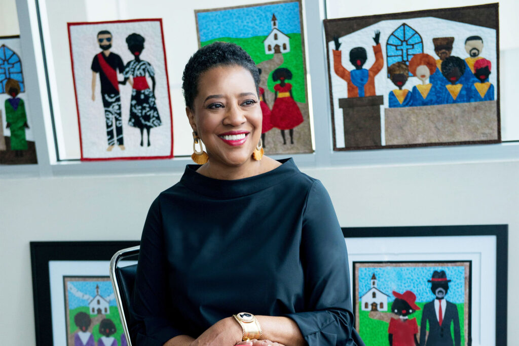 Photo: A black woman with a joyful smile sits in front of a wall of interesting art