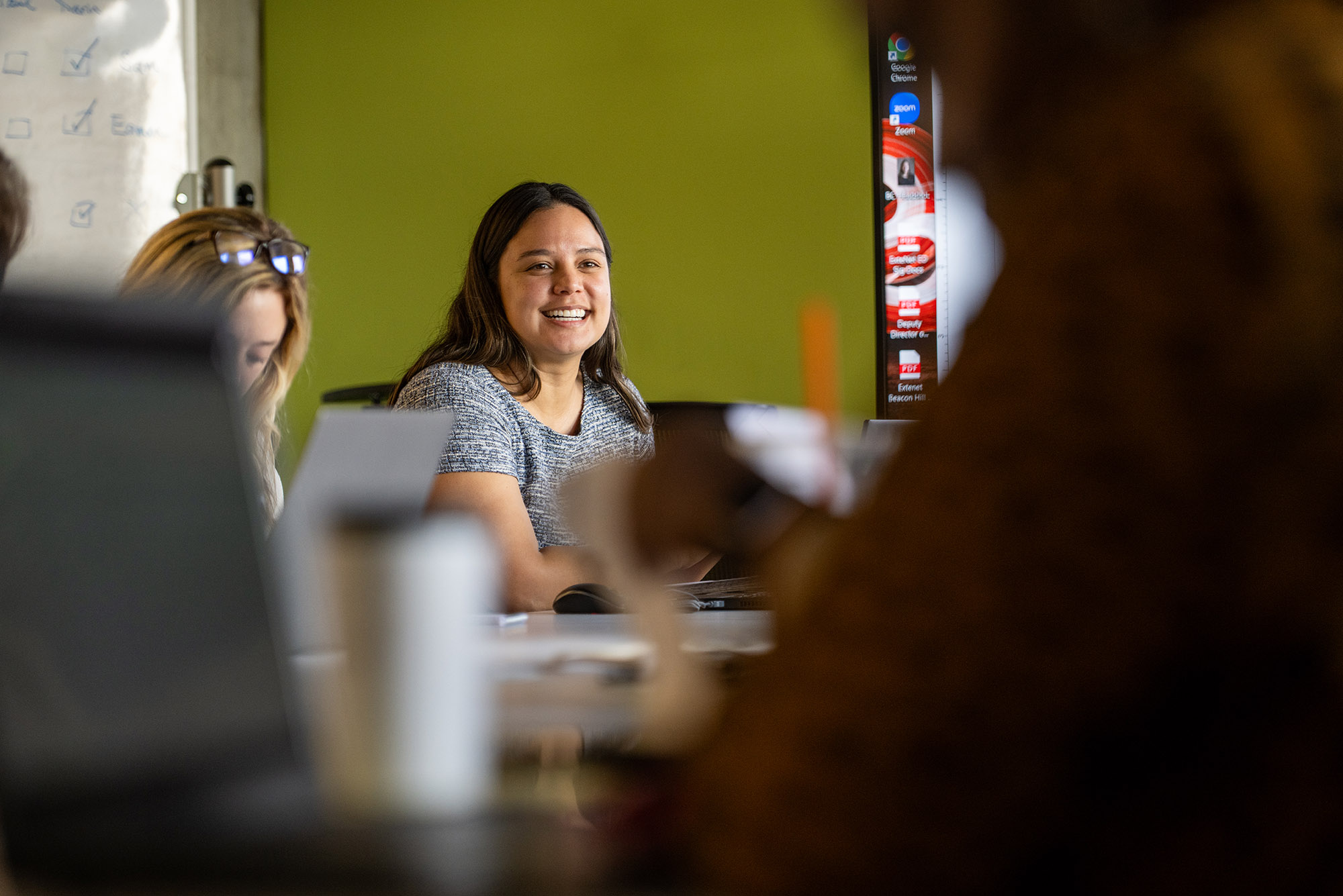 Photo: Beata Coloyan, a young woman, smiles at a meeting she holds. She wears a gray tshirt and shits around a table with her laptop in front of her.