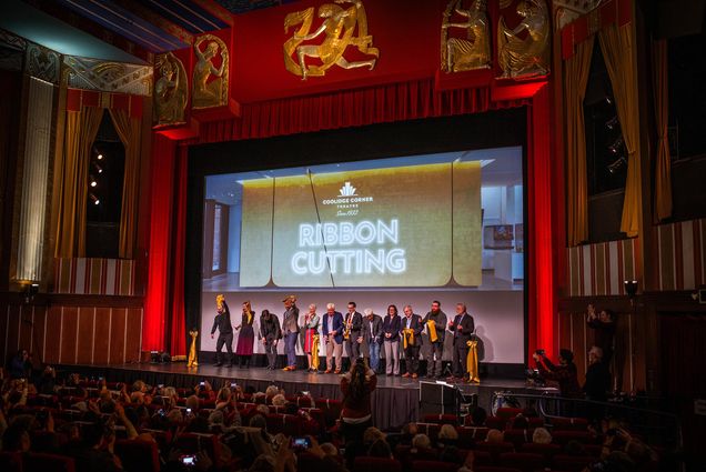 Photo: A picture of a stage with many people on it, all holding pieces of ribbon. There is a projection on the back of the stage that says "Coolidge Corner Theater, Since 1933, Ribbon Cutting"