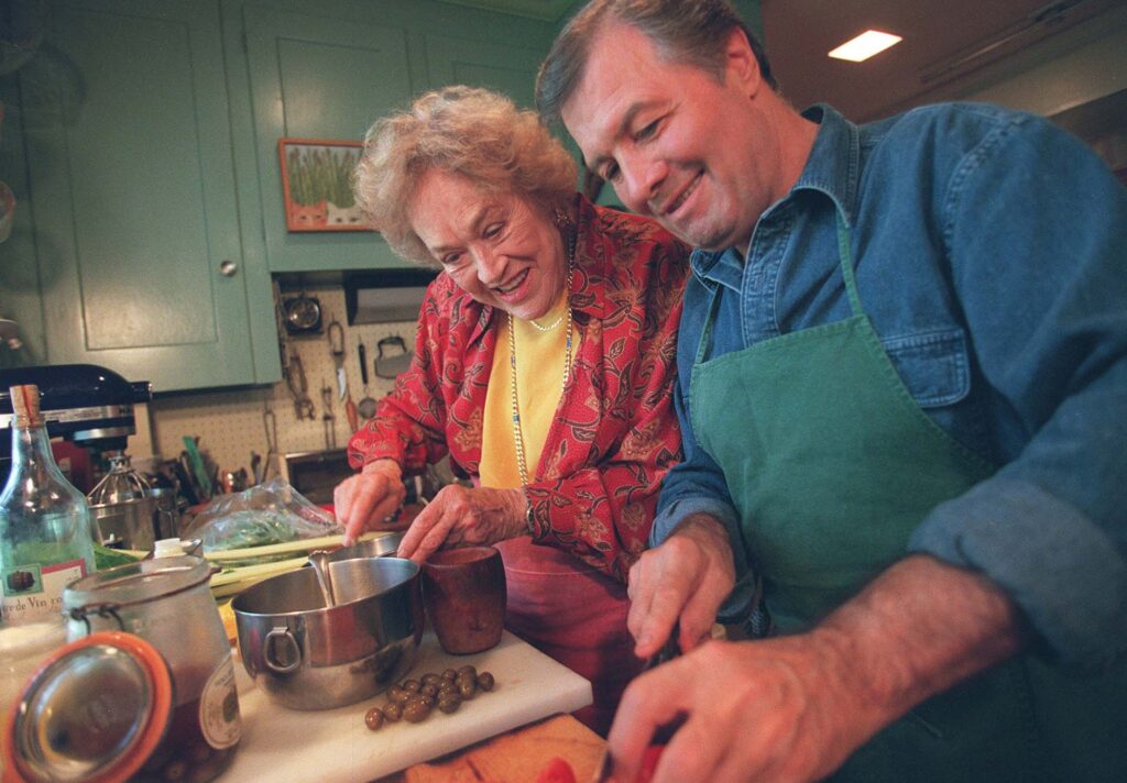 Photo: Julia Child and Jacques Pepin on their cooking show. Child wears a red button up with a yellow shirt and Pepin wears a denim shirt and a green apron. They smile as they prep their food.