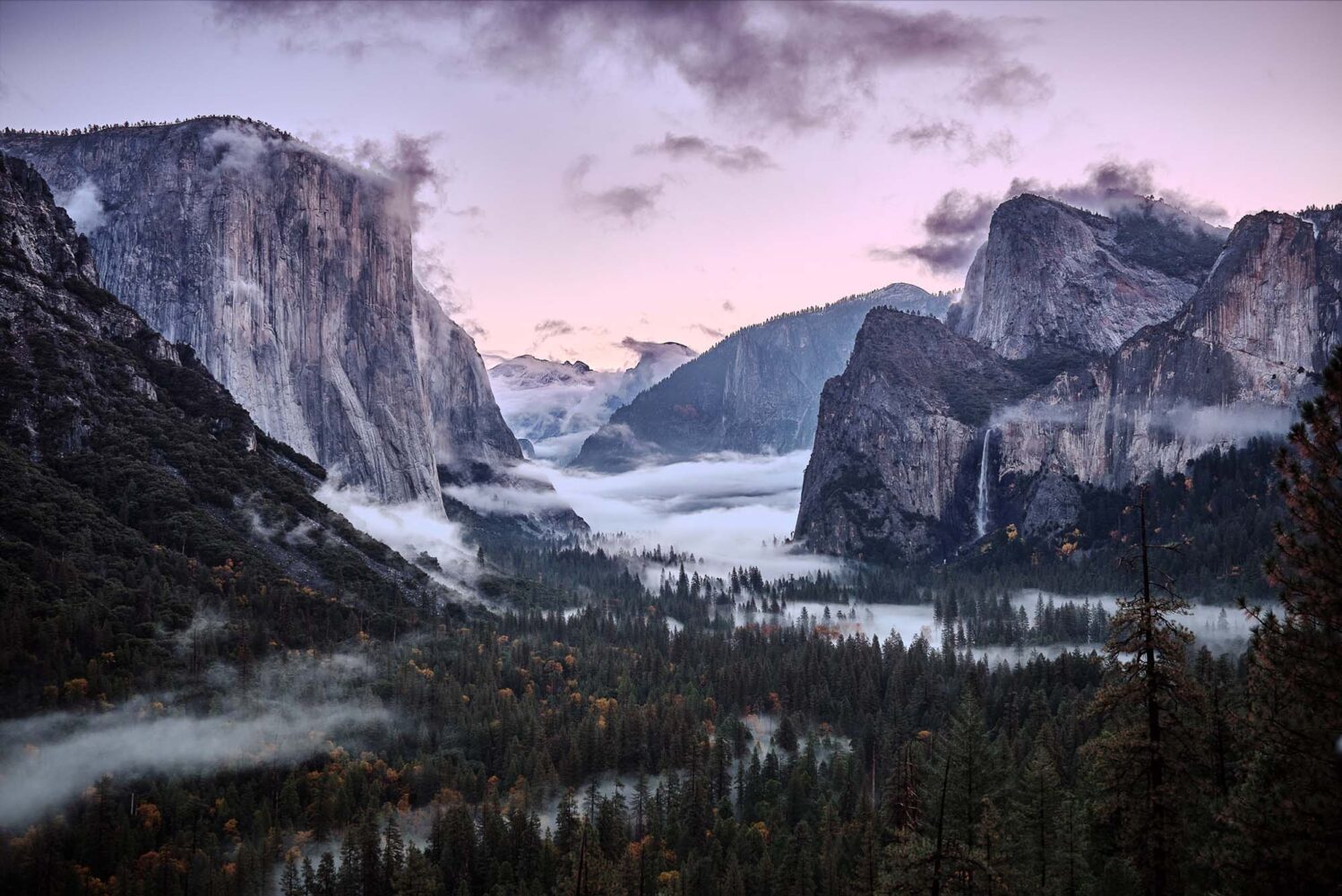 Photo: A wide shot of the mountain range at Yosemite National Park. The shot was taken at dawn, pink colors scatter across the sky while clouds hand low and skirt around the treeline.